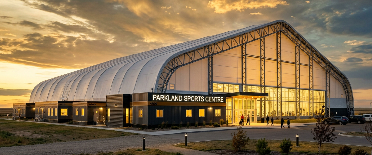 Aerial view of a modern soccer facility with two full-size 9v9 artificial turf fields side by side, each covered by large white double truss shelter structures, clean modular sports complex design, players training on one field and a game on the other, flat Alberta prairie landscape, dramatic sky with gold and deep blue tones, professional architectural rendering style